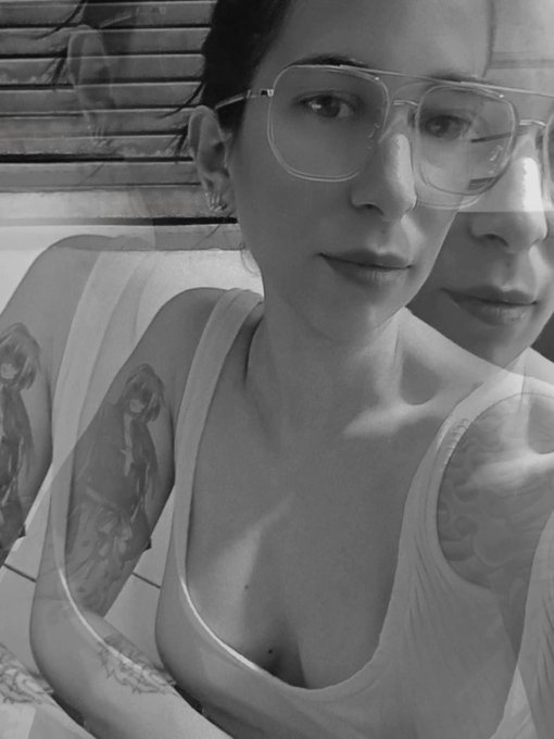 A black-and-white selfie shows a woman with glasses and anime character tattoos on her arm.