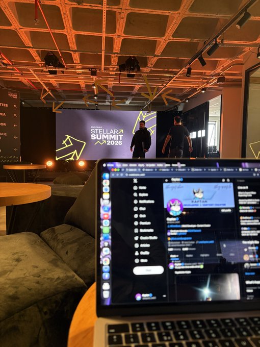 A laptop sits on a beanbag in the foreground displaying a chat window, while in the background a stage screen reads "Stellar Summit 2026" as attendees gather at the event—capturing the casual, behind-the-scenes "gm" morning vibe of the Stellar Builder Summit.