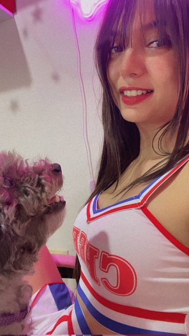 A selfie of a young woman with long brown hair wearing a white and red cheerleader-style outfit, smiling next to a small dog in a room with pink/purple neon lighting.