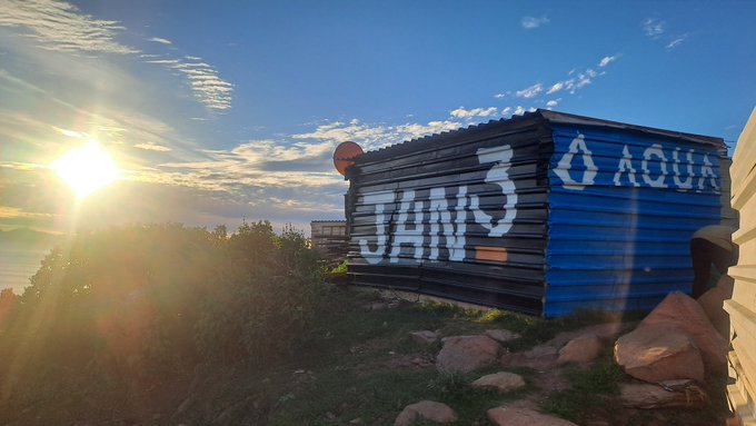 A corrugated metal structure displays the hand-painted text "JAN 3" and "AQUA" in white lettering against a blue background, with the sun rising brightly over a rocky, vegetated landscape. The "AQUA" signage directly references the @AquaBitcoin community center mentioned in the morning greeting, tying the scenic sunrise shot to the Bitcoin Ekasi initiative.