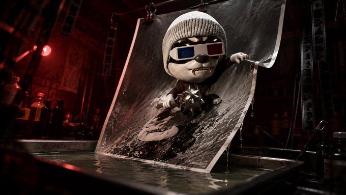 A creative photograph of a small character with 3D glasses and a beanie pulling a large black-and-white print out of a darkroom developing tray, with a mace-like object in the foreground.