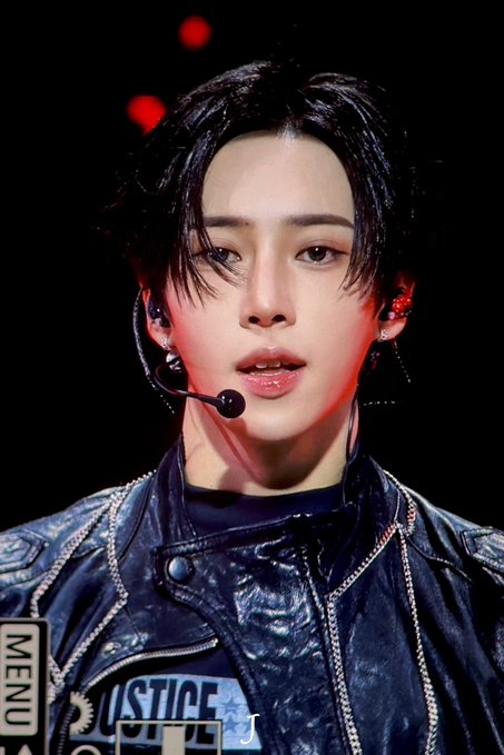 A performer with dark hair and a headset microphone performs on stage, wearing a distressed leather jacket featuring "JUSTICE" text on the front. The image relates to the hashtags by capturing a stage moment from the ALPHA DRIVE ONE (ALD1) world tour, with "MENU" and "J" text visible at the bottom corners likely indicating broadcast graphics.