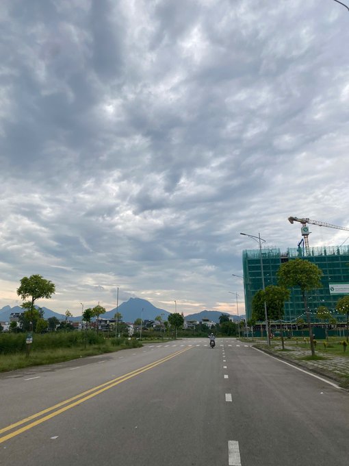 A serene road scene under a dramatic cloudy sky, with mountains in the background, a construction site with a crane on the right, and a lone motorcyclist riding down the street.