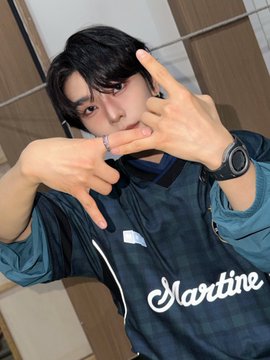 The image features a young man posing for a picture, wearing a black and blue shirt with white lettering that says "Martine". He has his hands up in front of him, making a peace sign gesture as he poses for the photo. The background is blurred, but it appears to be a room or studio setting where the photo was taken.