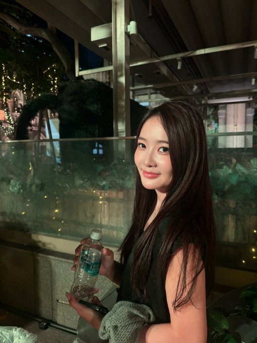 A young woman with long dark hair smiles while holding a bottled water, posing in what appears to be an outdoor evening venue with string lights and greenery in the background. The casual setting complements the post's mention of enjoying time at the BNB Chain event in Singapore, capturing a relaxed moment from the gathering.