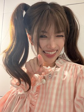 A cheerful young woman with pigtails and a pink bow smiles brightly while pointing at the camera, wearing a pink striped top and a heart necklace.