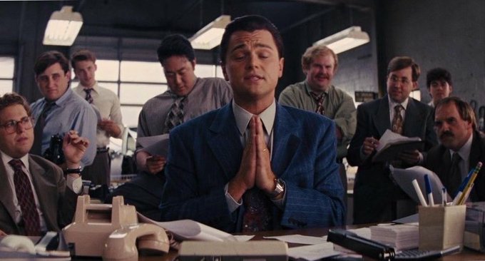 A scene from the movie 'The Wolf of Wall Street' showing Leonardo DiCaprio in a blue suit with hands clasped in prayer, surrounded by colleagues in an office.