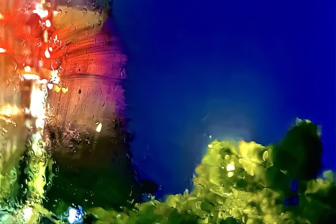 An abstract, impressionistic artwork features vibrant, blurred colors with deep blues, bright greens, and warm reds blending together in textured brushstrokes.