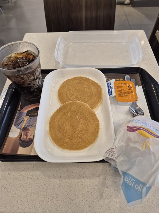 A McDonald's breakfast tray containing two pancakes, a hash brown in a wrapper, syrup, and a glass of iced soda on a speckled countertop.