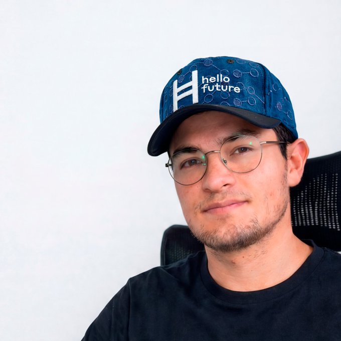 A young man with glasses and a beard wears a blue cap with the 'hello future' logo, sitting in a black chair and smiling at the camera.
