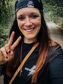 A selfie of a smiling woman wearing a black bandana and t-shirt with an orange strap, holding up a peace sign on a wooded path.