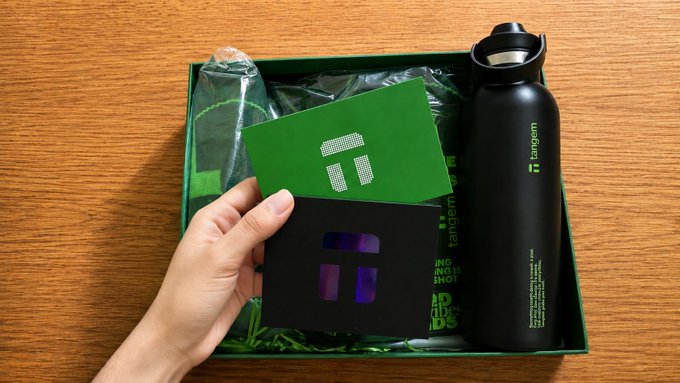 A hand holds up a Tangem card and a holographic "T" card from a green gift box containing a black Tangem-branded insulated water bottle, showcasing the crypto wallet company's branded merchandise. The "TANGEM" logo and "HOT AND NOT SHOT" text visible on the packaging align with the post's theme of receiving cool gear from the Tangem team for keeping crypto secure.
