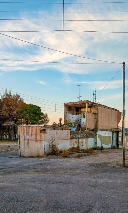 An old, weathered building with a tile roof stands against a cloudy blue sky, with power lines crossing overhead and a dirt area in the foreground.