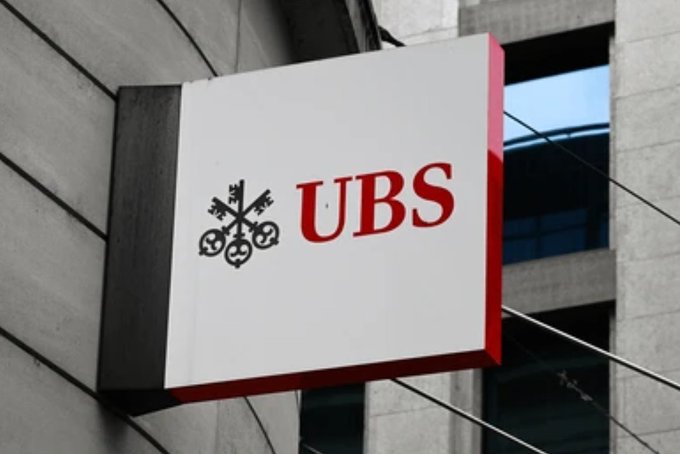 A UBS bank sign is mounted on the exterior of a building, featuring the company's red logo and crossed keys emblem. The image relates to the post by visually representing UBS, the Swiss bank highlighted for significantly increasing its spot Bitcoin ETF holdings by 230% to $90 million.