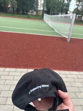 A hand holds up a black baseball cap embroidered with the "Expecting More" Extended logo in front of a tennis court, showcasing the merch received from the Extended crypto community to celebrate membership in the group.