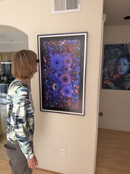 A woman with short blonde hair and glasses admires a vibrant framed artwork featuring purple daisies against a floral background, with another colorful portrait visible on the adjacent wall. The scene captures her appreciation for physical NFT-linked art, showcasing the real-world appeal of blockchain-based creations.
