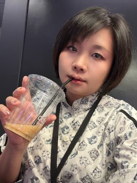 A young woman with short dark hair drinks from a clear plastic cup containing an orange beverage through a black straw, wearing a white shirt with a black pattern.
