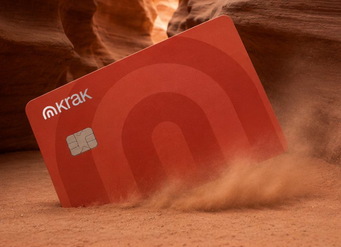 A red Krak debit card (branded with Kraken's payments app logo) is partially buried in sand with dust swirling around it, visually emphasizing the card's U.S. launch.