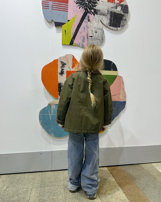 A young girl with a long blonde braid and an olive green jacket stands in a gallery, looking at a large colorful abstract artwork mounted on a white wall.