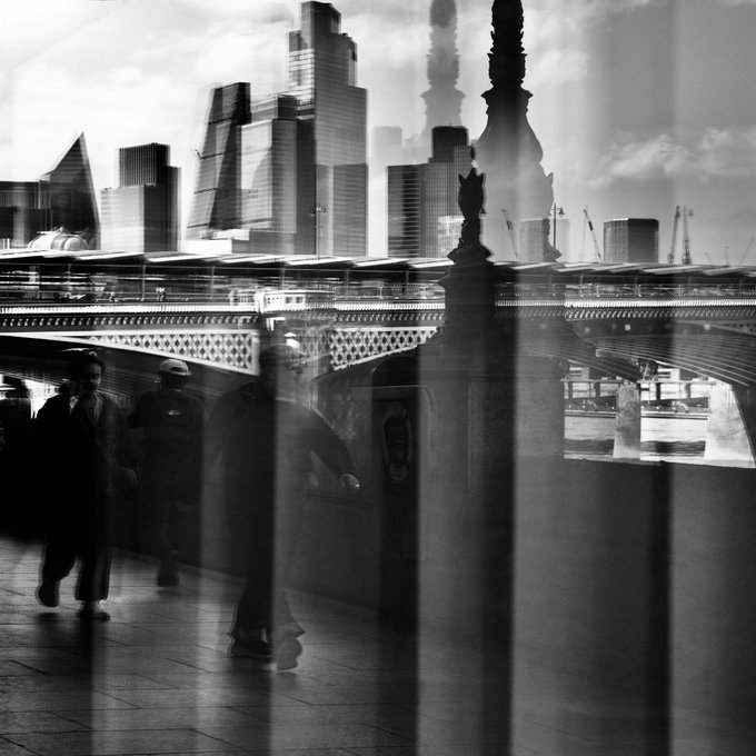 The image shows a cityscape with tall buildings, including a bridge that spans across the scene. The photo appears to be taken from a low angle, giving an interesting perspective of the city's skyline. A group of people can be seen walking on the sidewalk in front of these buildings, adding life and movement to the scene. The image also contains text elements such as "Parallel4 1/1 Res 50 $XTZ Live auction on @artcommissiontz objkt.com/tokens/KT1HiNN". This suggests that this photo might be part of an online auction or a digital art piece, where viewers can interact with the image and learn more about its context.