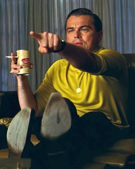 Leonardo DiCaprio as Jordan Belfort in 'The Wolf of Wall Street' pointing at the camera while holding a can, dressed in a yellow turtleneck with his feet propped up.