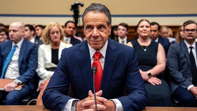The image shows a man sitting at a desk wearing a suit and tie, holding a microphone, likely speaking into it during a press conference or event. The man appears to be addressing an audience of people who are seated behind him. The text "LATEST: Former New York Gov. Andrew Cuomo, now co-chair of an OKX-ICE crypto venture, urged Congress to pass the CLARITY Act quickly", and "Focus on what is in the image and how it relates to the post" suggest that this man might be discussing a recent development or announcement related to cryptocurrency regulation or blockchain technology.