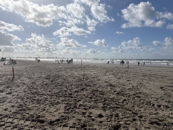 A wide sandy beach stretches toward the sea under a bright blue sky filled with scattered clouds, with small groups of people walking along the shoreline and tire tracks visible in the sand.