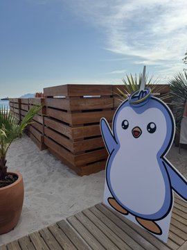 A cartoon penguin cutout with a crown stands on a wooden deck beside a wooden planter, with palm plants and the ocean visible in the background.