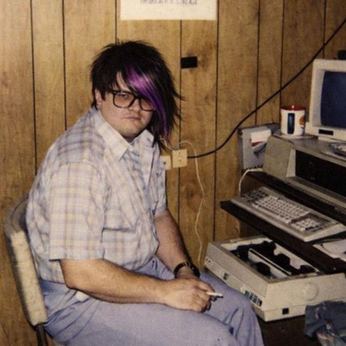 A young man with emo-style purple-streaked hair and black glasses sits at a desk with a vintage computer, smoking a cigarette in a wood-paneled room.