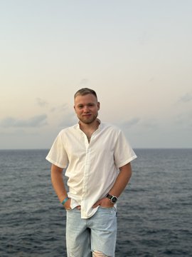 A man with short blonde hair stands outdoors by the sea at dusk, wearing a white short-sleeved shirt and light blue denim shorts, with a watch on his left wrist.