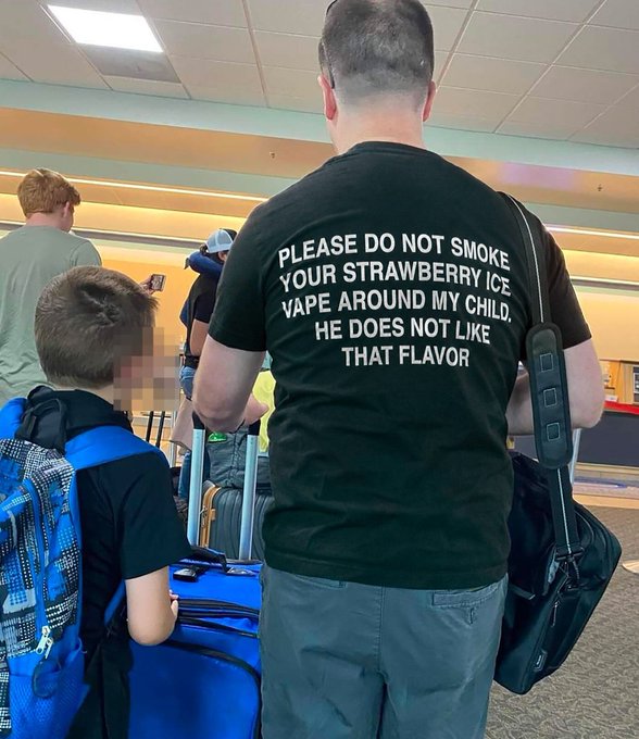 A man at an airport wears a black t-shirt with white text reading 'PLEASE DO NOT SMOKE YOUR STRAWBERRY ICE VAPE AROUND MY CHILD, HE DOES NOT LIKE THAT FLAVOR,' standing next to a child with a backpack and luggage.