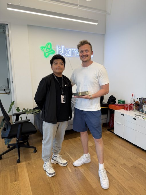 Two men pose inside the Nansen HQ in Singapore, with the illuminated "Nan..." logo visible on the wall behind them; one holds what appears to be the NVIDIA DGX Spark device mentioned in the caption. The image captures the moment of delivering the AI hardware during the visit.
