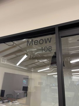 A glass office door labeled 'Meow 108' reveals a modern, open-plan office space with desks, chairs, and overhead lighting visible inside.