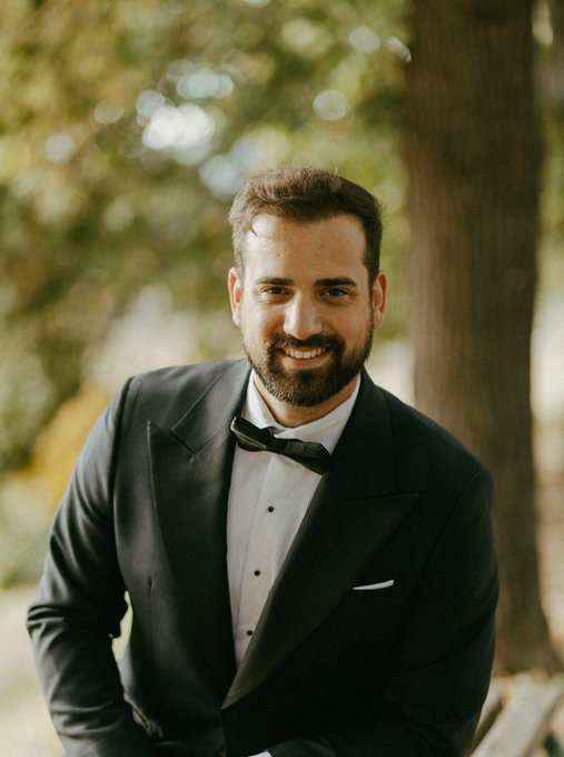 A portrait of a smiling bearded man in a classic black tuxedo with a bow tie, photographed outdoors with a soft, blurred background.