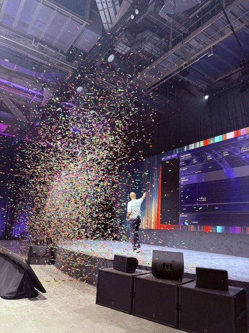 A person on stage celebrates as colorful confetti explodes into the air during what appears to be a concert or event, with large speakers visible in the foreground.