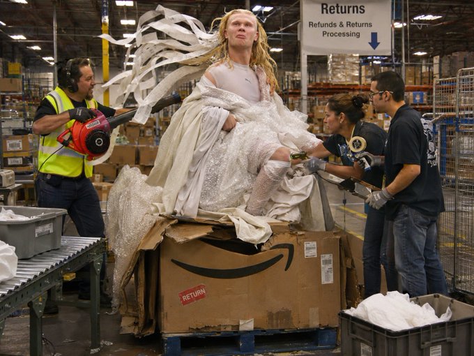 A surreal composite image showing a woman in an elaborate white gown emerging from a cardboard Amazon box in a warehouse returns processing area, while workers in safety vests handle packages nearby.