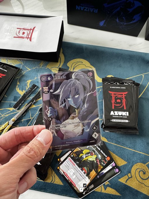A hand holds an Azuki trading card game card featuring the character Raizan, alongside booster packs labeled 'AZUKI TRADING CARD GAME' and a playmat.