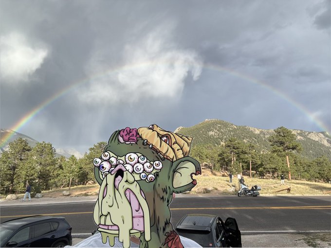 A surreal cartoon scene shows a green monster character with an exposed brain and multiple eyes, set against a backdrop of mountains and a full rainbow.