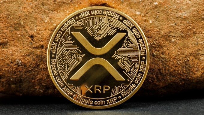 The image features a gold coin with an XRP symbol on it. The coin has been placed on top of a rock, which adds a rustic touch to the scene. The text accompanying the image reads "XRP is going to flip $BTC within 90-days from this date. Before or on July 18, 2026 $XRP will be number 1 crypto." This message suggests that the XRP coin has been minted and is now being released into circulation with a specific timeframe in mind.