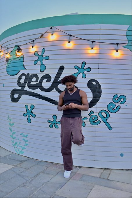 A man in a tank top and pants stands on one leg in front of a white wall featuring 'geck crêpes' graffiti and string lights, looking at his phone.