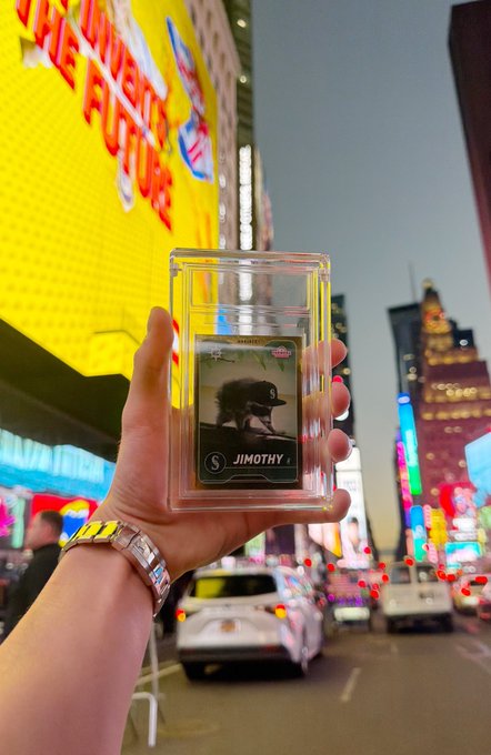 A hand holds up a $Jimothy trading card in Times Square, featuring the token's mascot—a cap-wearing figure—with the address "VJ6SX...F7y2," promoting a giveaway of 1,000,000 $Jimothy tokens to commenters sharing their Solana public address.