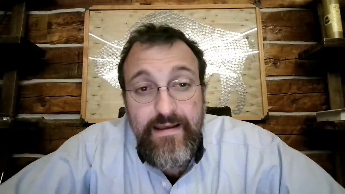 The image shows a man wearing glasses, sitting at a desk with his hands on his lap. He appears to be looking directly into the camera while talking. There are several books scattered around him, suggesting that he might be engaged in an intellectual discussion or research. The presence of these books and the man's focused expression indicate that this could be a significant moment in his work or study.