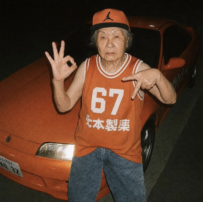 An elderly woman wearing an orange '67' tank top and orange Jordan cap makes hand gestures while standing in front of an orange sports car at night.