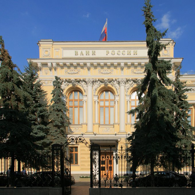 The image shows the ornate yellow neoclassical facade of the Bank of Russia ("БАНК РОССИИ") headquarters with its flag flying atop, framed by tall evergreen trees and an iron gate. This central bank building directly connects to the news that Russia's monetary authority has flagged cryptocurrencies and stablecoins as threats to its sovereignty and the ruble.
