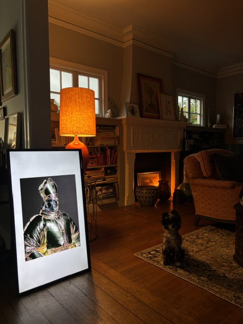 A warmly lit living room with a small dog looking toward a framed artwork on the floor, an orange table lamp, fireplace, armchair, and bookshelves.