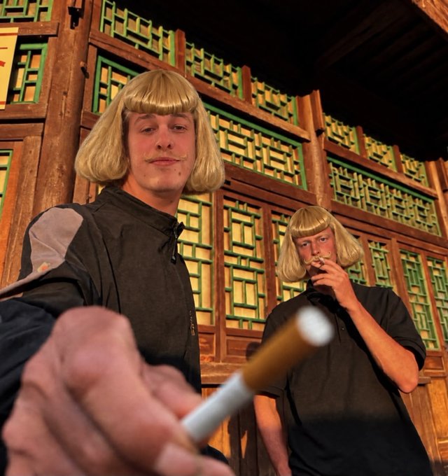 Two people wearing blonde bob wigs and dark clothing pose for a photo in front of traditional Chinese wooden lattice windows, with one holding out a cigarette.