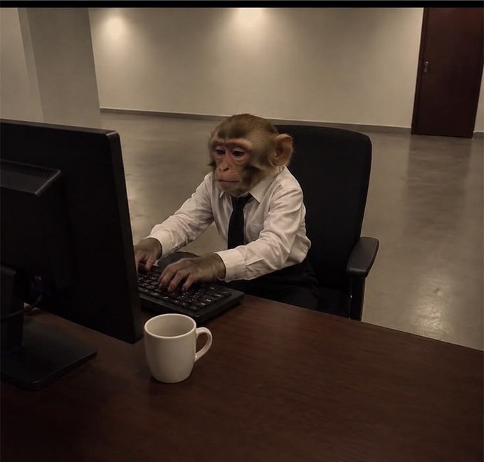 A monkey dressed in a white shirt and dark tie sits at an office desk typing on a keyboard in front of a computer monitor, with a white mug nearby.