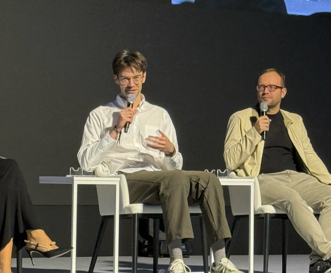 Two men wearing casual button-up shirts sit on white stools on a stage, each holding a microphone as they appear to be speaking during a panel discussion or Q&A session.