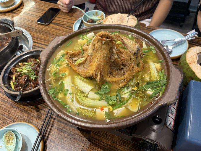 A large hot pot of fish soup with vegetables and fried fish, served on a portable burner alongside side dishes at a wooden table.