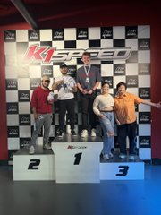 A group of men pose for a photo on a podium (#1, #2, #3) in front of a K1 SPEED banner, with one man holding a trophy.